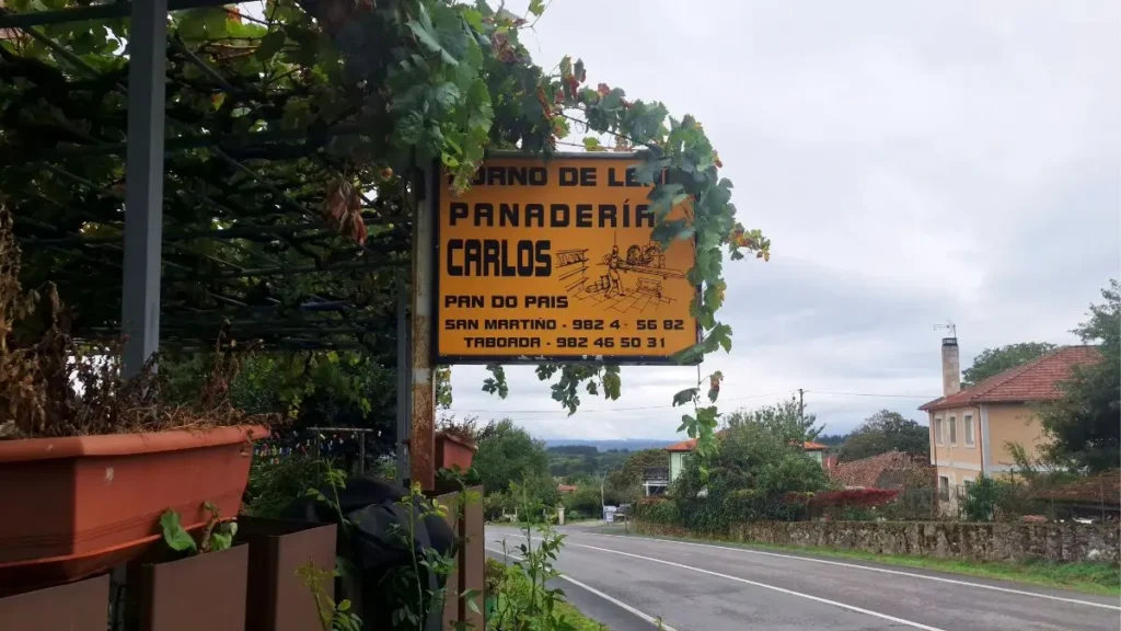 Roadside sign reading Forno de leña, Panadería Carlos, Pan do país, under a vine pergola